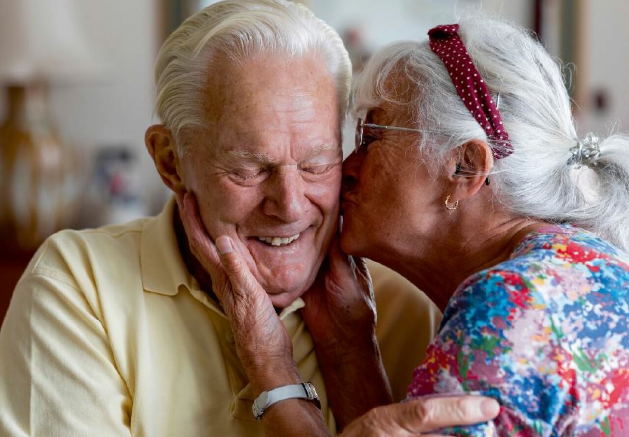 Twee oude mensen zitten en lachen. De een geeft de ander een zoen op de wang, ze lachen en kijken gelukkig.