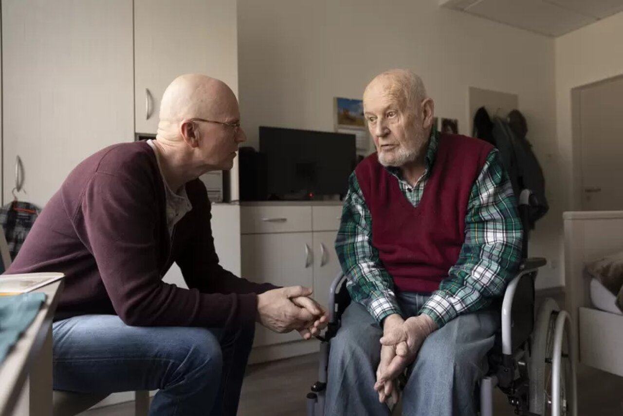 Een oude man en een verzorger zitten samen in een kamer en hebben een gesprek, foto uit online artikel Het Parool.