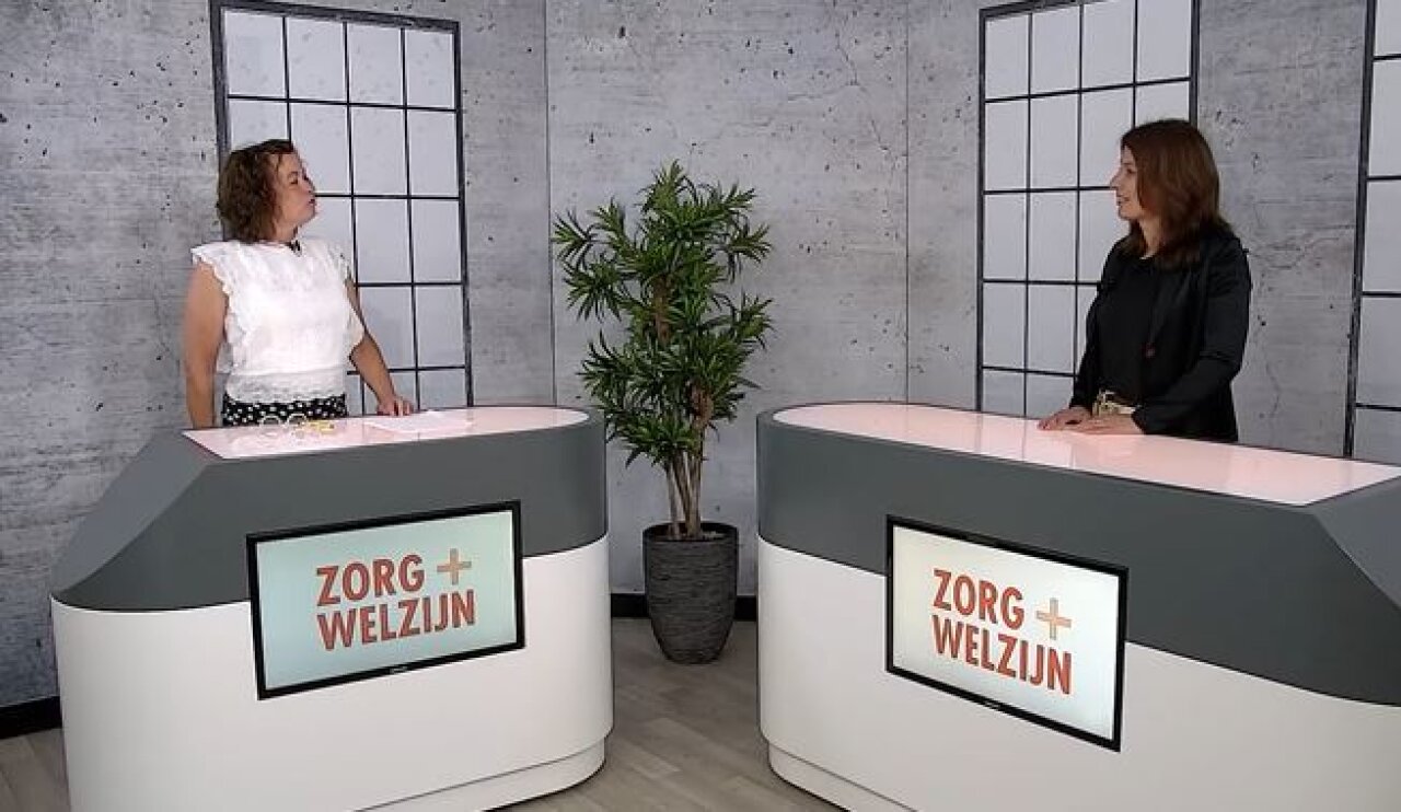 Een presentatrice en Anne Mei The staan elk achter een desk en praten met elkaar.